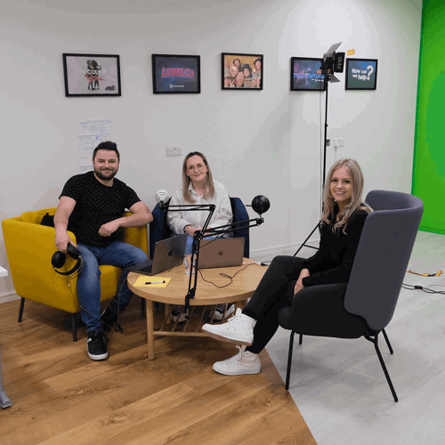 Level Up Podcast Recording: Two ClassForKids team members and Lauren, the Level Up contest winner, sit around a podcast setup in a bright studio. Two women and one man smile toward the camera, seated with laptops and microphones. Framed posters from ClassForKids campaigns hang on the wall behind them.