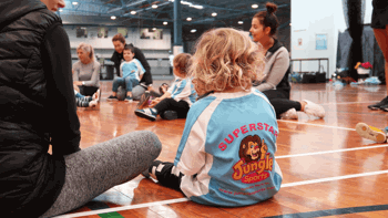 A young child with curly blonde hair sits on a wooden indoor sports court, wearing a blue Jungle Sports jersey that says “Superstar” with a cartoon lion logo. Around them, other children and adults sit in a circle, engaging in a playful, structured session.
