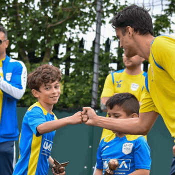 Coach fist bumping one of the young team mates (a boy in a blue football strip). Another coach is in the background clapping and there are other members of the team visible.