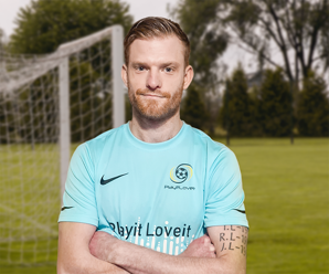 A football coach stands confidently with arms crossed on a grassy pitch in front of a goal. He wears a light blue “Play it Love it” branded jersey.