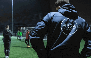 Two coaches wearing dark jackets with “AR26 Charity” printed on the back observe a group of young players during a football training session on a floodlit pitch.