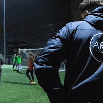 Two coaches wearing dark jackets with “AR26 Charity” printed on the back observe a group of young players during a football training session on a floodlit pitch.