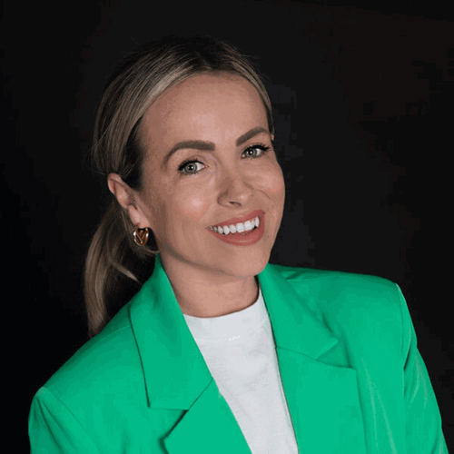A woman with blonde hair tied back and wearing a vibrant green blazer smiles confidently at the camera. The background is dark, making her outfit and bright expression pop.