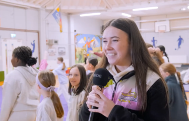 Brunette female gymnastics coach holding a microphone and smiling with gymnastics class behind her