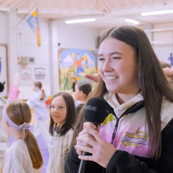 Brunette female gymnastics coach holding a microphone and smiling with gymnastics class behind her