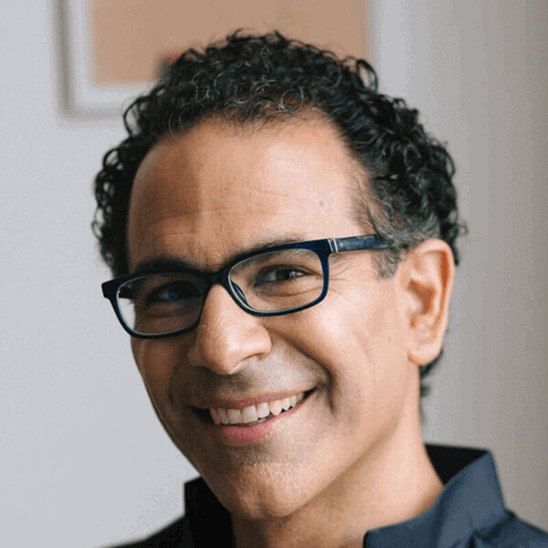 A smiling man with short curly hair and glasses, wearing a dark shirt, stands in a softly lit indoor space. He appears relaxed and confident, facing the camera.