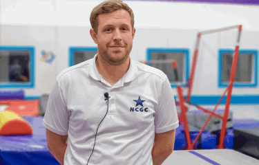 A male coach wearing a white polo shirt with the NCGC (Nottingham City Gymnastics Club) logo stands in a bright, colourful gymnastics facility. Behind him, padded equipment and uneven bars are visible.