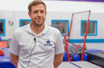 A male coach wearing a white polo shirt with the NCGC (Nottingham City Gymnastics Club) logo stands in a bright, colourful gymnastics facility. Behind him, padded equipment and uneven bars are visible.