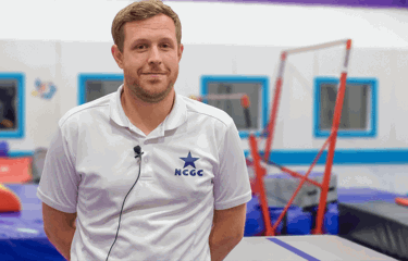 A male coach wearing a white polo shirt with the NCGC (Nottingham City Gymnastics Club) logo stands in a bright, colourful gymnastics facility. Behind him, padded equipment and uneven bars are visible.