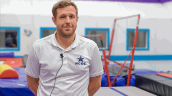 A male coach wearing a white polo shirt with the NCGC (Nottingham City Gymnastics Club) logo stands in a bright, colourful gymnastics facility. Behind him, padded equipment and uneven bars are visible.
