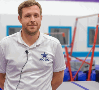A male coach wearing a white polo shirt with the NCGC (Nottingham City Gymnastics Club) logo stands in a bright, colourful gymnastics facility. Behind him, padded equipment and uneven bars are visible.