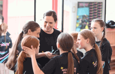 A smiling dance teacher chats warmly with a group of young dancers, who are all wearing matching Nicole O'Donnell Dance Academy t-shirts.