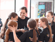 A smiling dance teacher chats warmly with a group of young dancers, who are all wearing matching Nicole O'Donnell Dance Academy t-shirts.