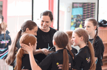 A smiling dance teacher chats warmly with a group of young dancers, who are all wearing matching Nicole O'Donnell Dance Academy t-shirts.