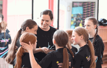 A smiling dance teacher chats warmly with a group of young dancers, who are all wearing matching Nicole O'Donnell Dance Academy t-shirts.