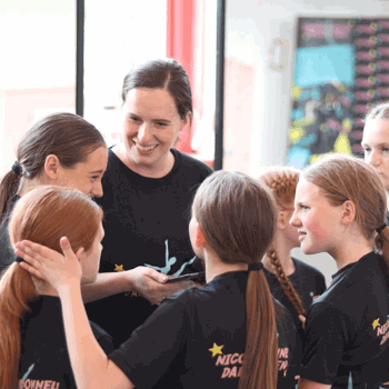 A smiling dance teacher chats warmly with a group of young dancers, who are all wearing matching Nicole O'Donnell Dance Academy t-shirts.