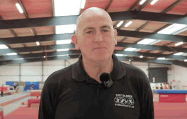 A man in a black polo shirt with the East Kilbride Gymnastics Club logo stands inside a large gymnastics facility. Colourful equipment and athletes training in the background.
