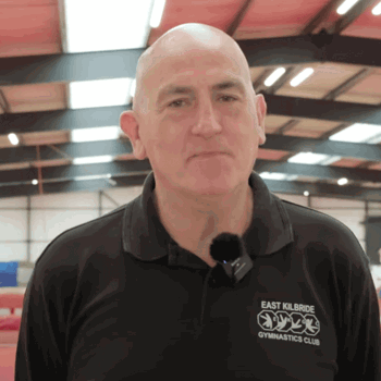A man in a black polo shirt with the East Kilbride Gymnastics Club logo stands inside a large gymnastics facility. Colourful equipment and athletes training in the background.