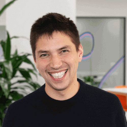 A man with short dark hair and a wide smile stands in a bright office, wearing a dark jumper. The background includes green plants and a blurred workspace, creating a cheerful and open atmosphere.