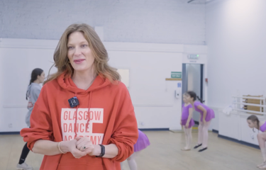 Blonde woman wearing an orange Glasgow Dance Academy hoodie with dancers in the background