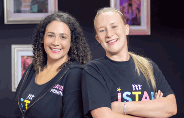 Two smiling women stand confidently back-to-back, wearing J Star Academy branded clothing.