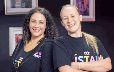 Two smiling women stand confidently back-to-back, wearing J Star Academy branded clothing.