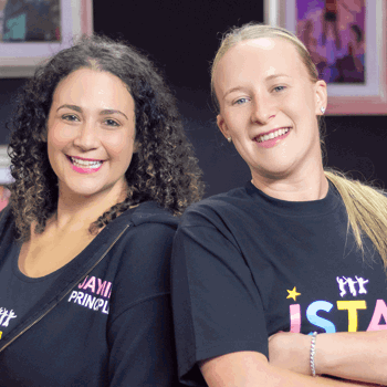 Two smiling women stand confidently back-to-back, wearing J Star Academy branded clothing.
