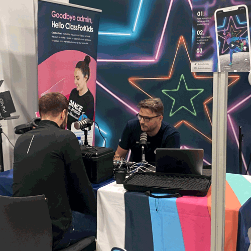A man wearing glasses sits at a table with podcasting equipment and a laptop, speaking into a microphone. Across from him, another man listens intently. Behind them is a vibrant neon star backdrop and a banner that reads “Goodbye admin, Hello ClassForKids.”
