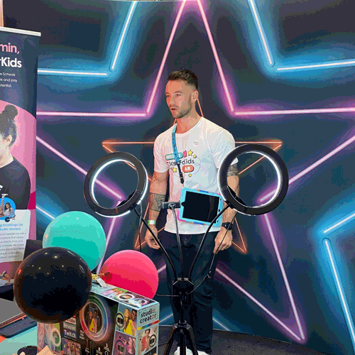 A ClassForKids team member stands in front of a vibrant neon star backdrop, wearing a branded t-shirt and lanyard. In front of him is a ring light setup with a tablet and colourful balloons, creating a fun, interactive booth experience.