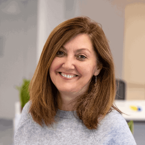A woman with long brown hair smiles warmly at the camera in a bright office space. She’s wearing a soft grey jumper and earrings, giving off a friendly, approachable vibe.