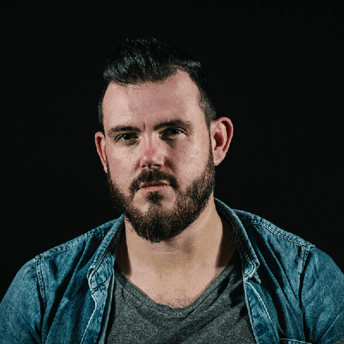 A man with a serious expression, trimmed beard and short hair, wears a denim shirt over a grey tee, posing against a dark background.