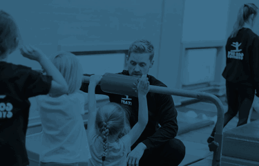 A gymnastics coach kneels beside a low bar, guiding a young child as they prepare to swing.