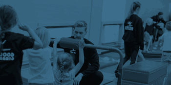 A gymnastics coach kneels beside a low bar, guiding a young child as they prepare to swing.