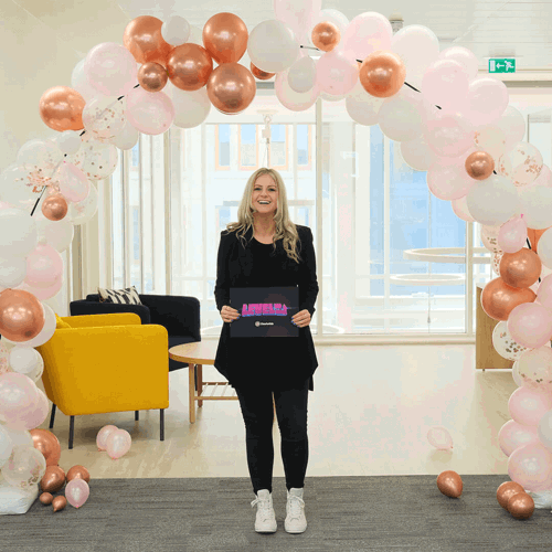 A woman stands smiling beneath a balloon arch made of pink, white and rose gold balloons in a modern office space. She holds a sign that says “Level Up” and wears all black with white trainers.